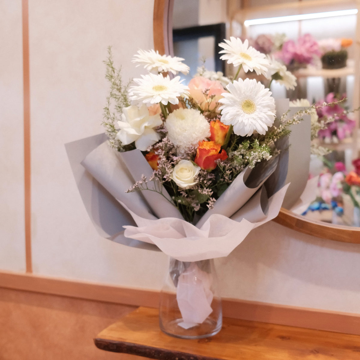 Cherished bouquet with white gerbera daisies, roses, and orange spray roses wrapped in paper, displayed in a clear glass vase