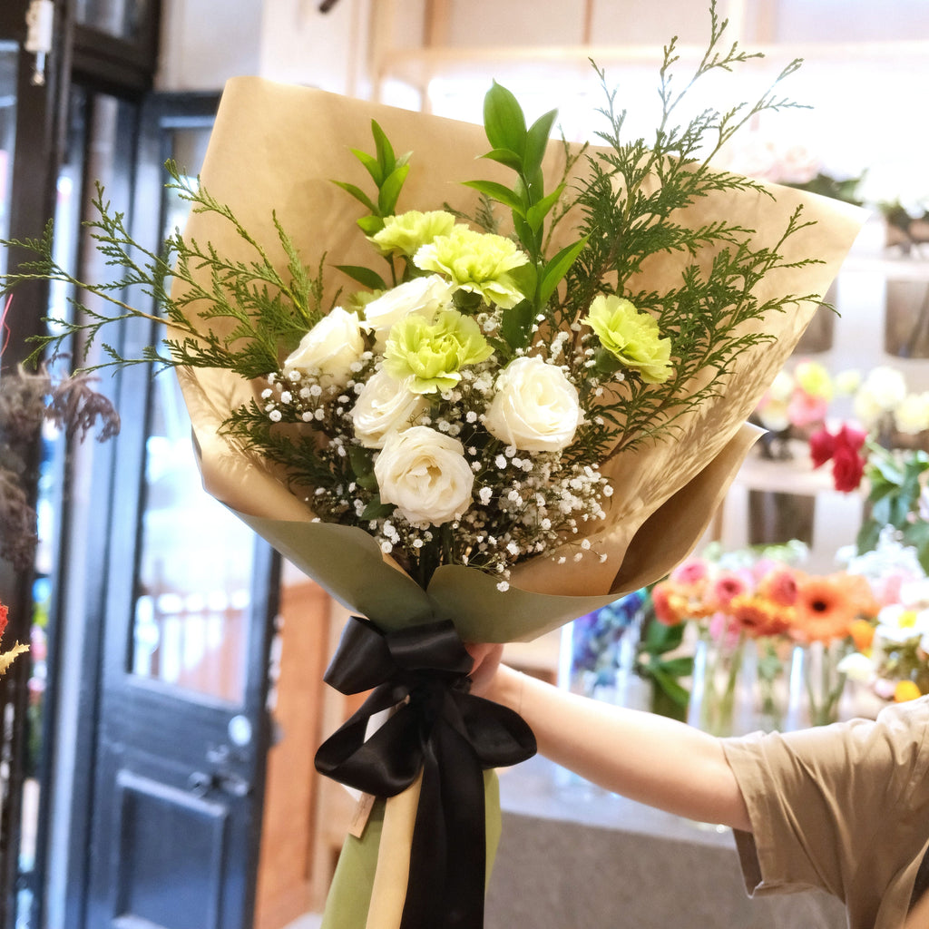 Florist holding the Gentle Remembrance white rose and sage carnation sympathy bouquet, wrapped in kraft paper with black ribbon