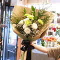 Florist holding the Gentle Remembrance white rose and sage carnation sympathy bouquet, wrapped in kraft paper with black ribbon