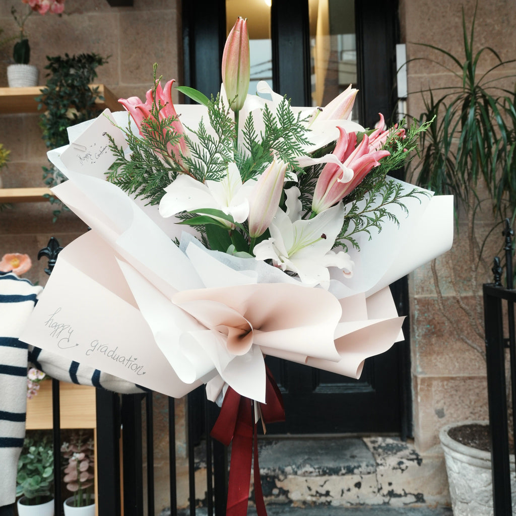 Mirai Grace pink and white lily graduation bouquet with "happy graduation" wrapping, outside a Sydney sandstone shopfront