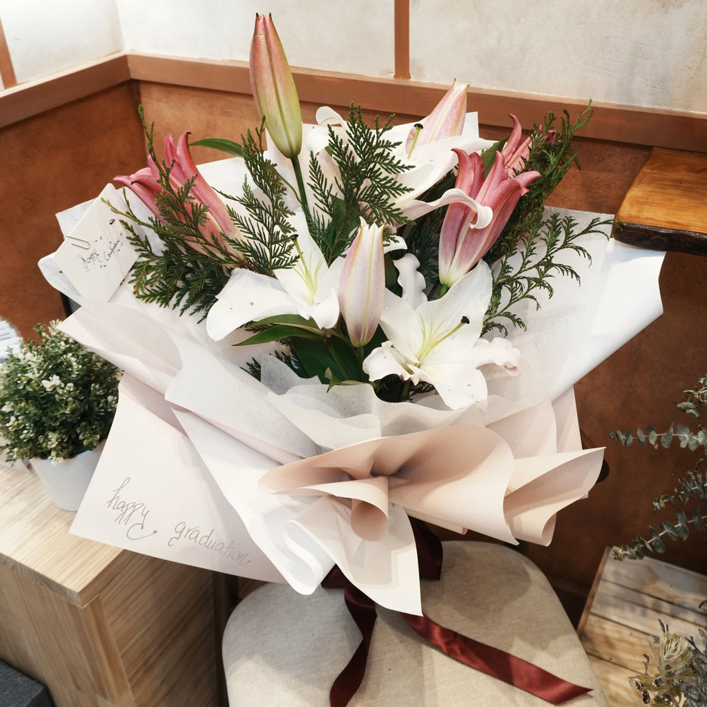 Mirai Grace graduation bouquet of white and pink Oriental lilies with cedar greenery, wrapped in white and blush paper with a burgundy ribbon