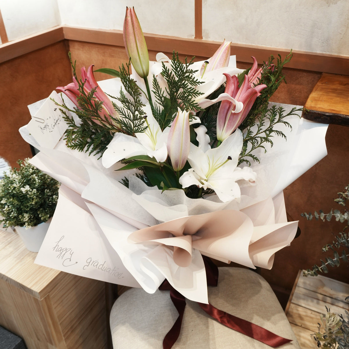 Mirai Grace graduation bouquet of white and pink Oriental lilies with cedar greenery, wrapped in white and blush paper with a burgundy ribbon