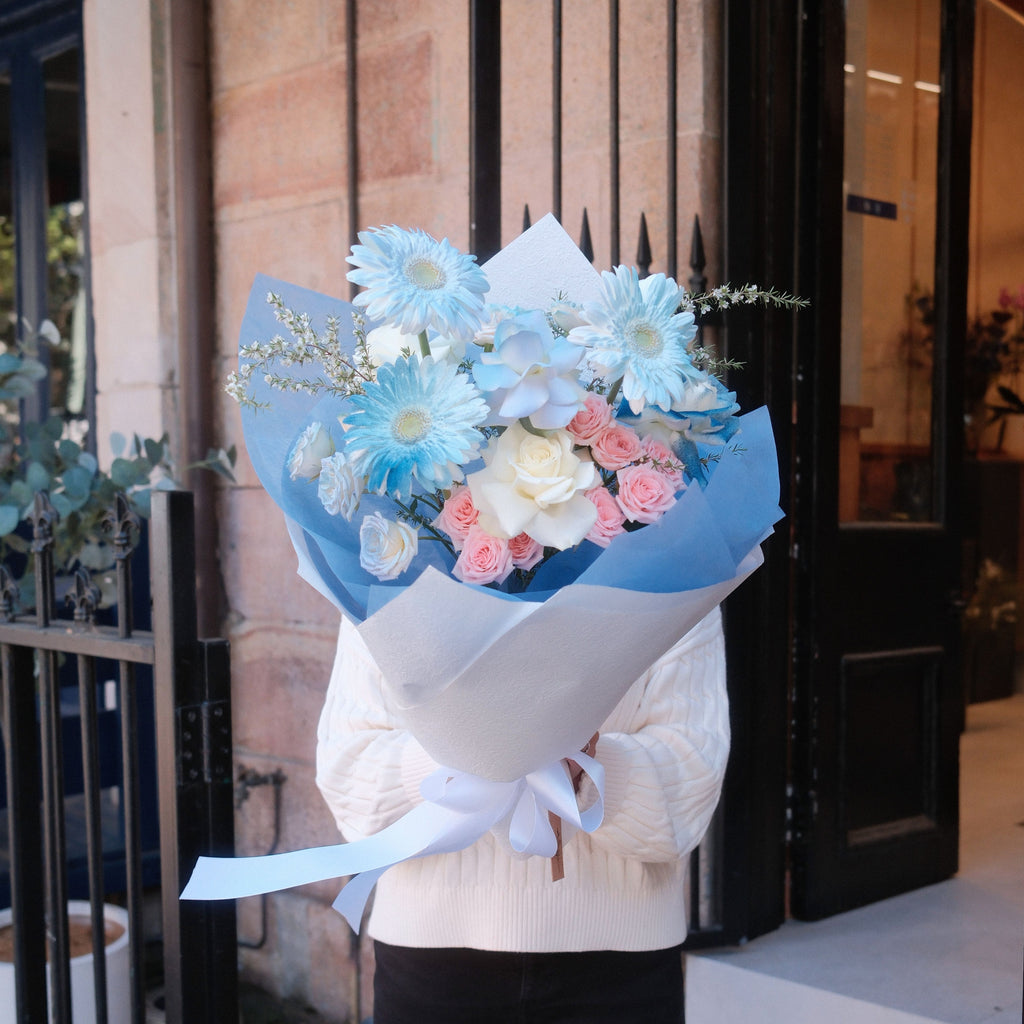 Sky Muse bouquet with blue gerbera daisies, white roses, blush spray roses, and white gardenia in blue and white wrapping paper.